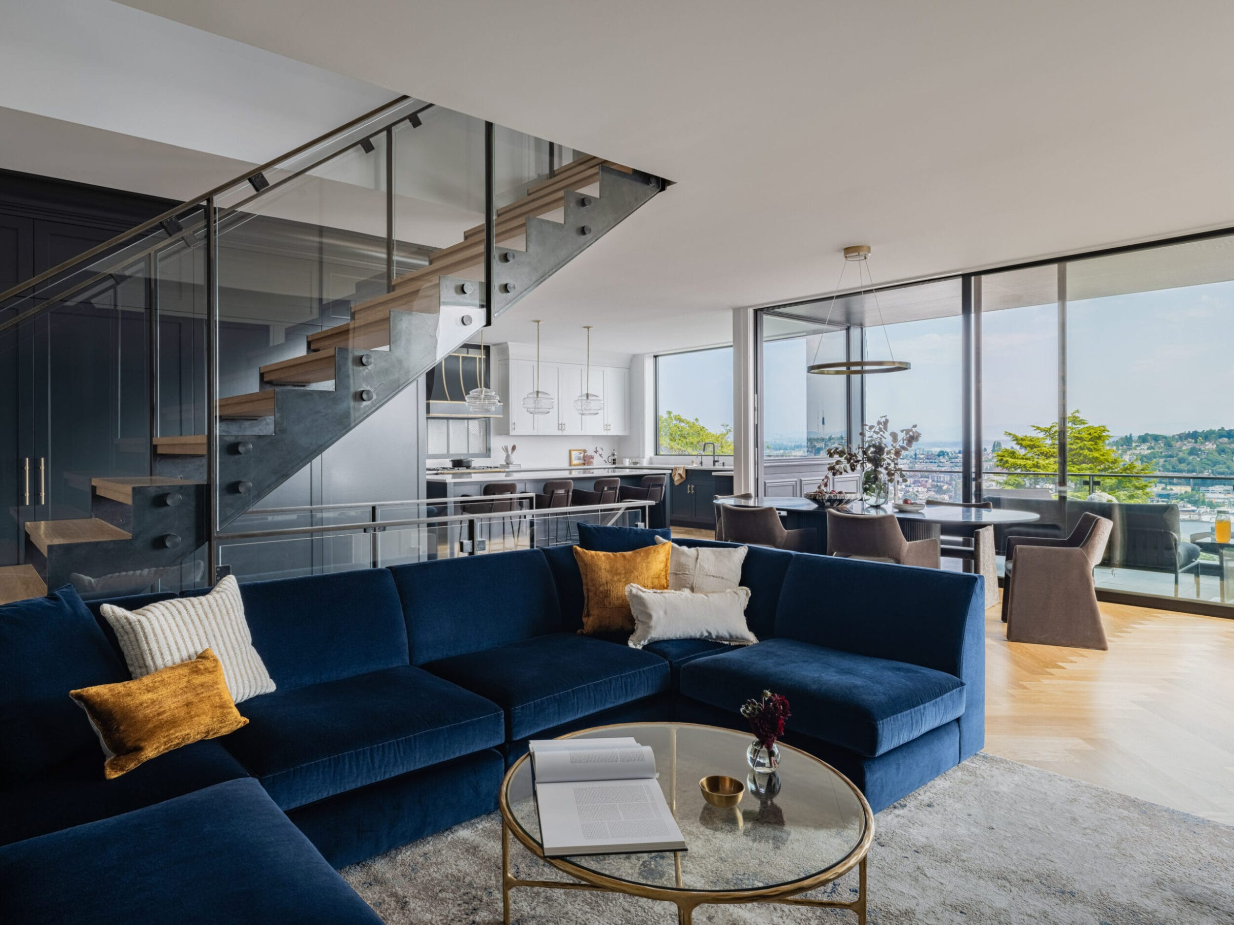 A modern living room with a blue sectional sofa, glass coffee table, and a staircase. Dining area and kitchen are visible in the background, with large windows overlooking a cityscape.