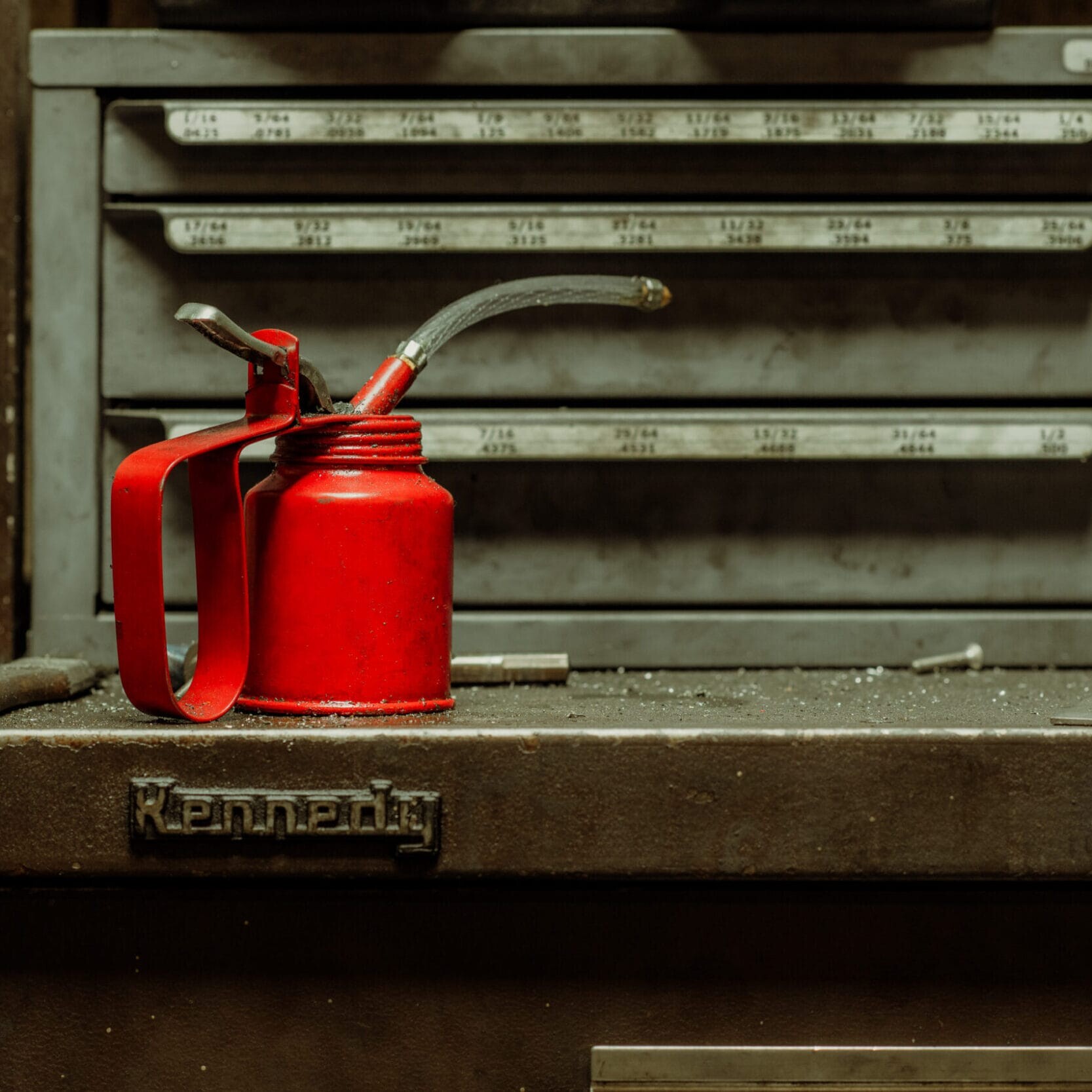 A red oil can with a curved spout sits on a dusty metal toolbox at Dyna Metal Shop Seattle, in front of a gray cabinet with labeled drawers.