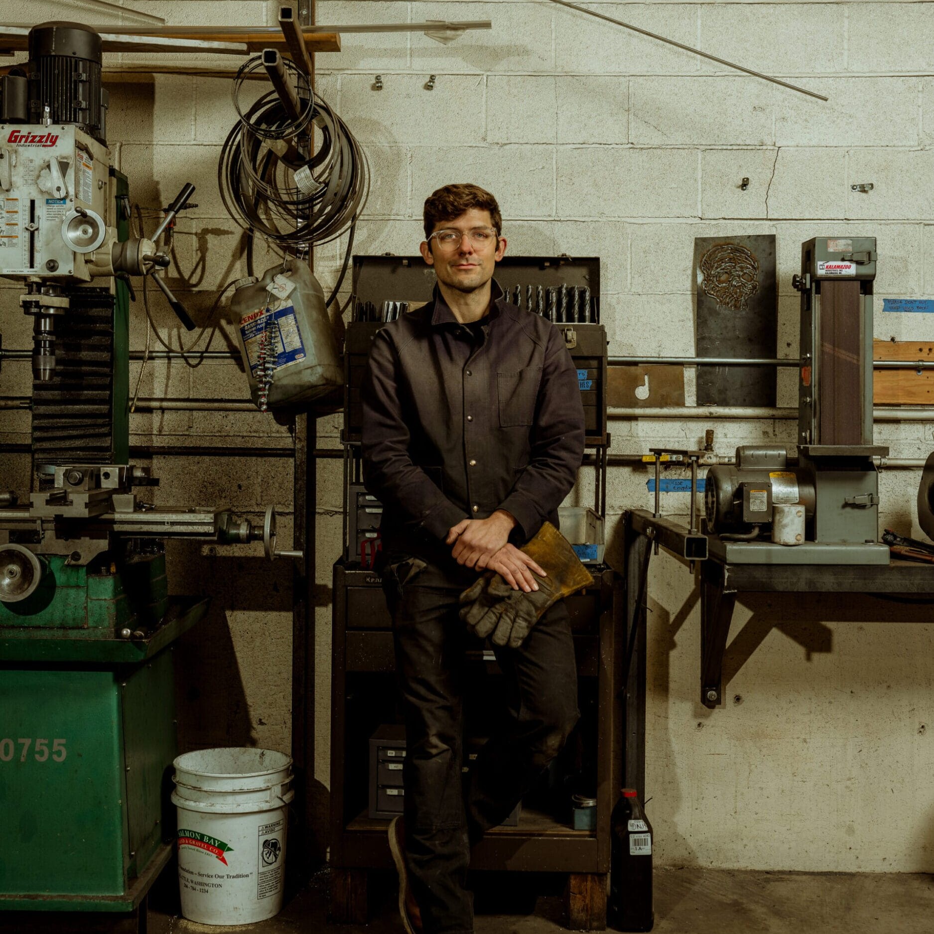 A person wearing dark work clothes and gloves stands in the Dyna Metal Shop Seattle workshop, surrounded by industrial tools and equipment against a cinder block wall.