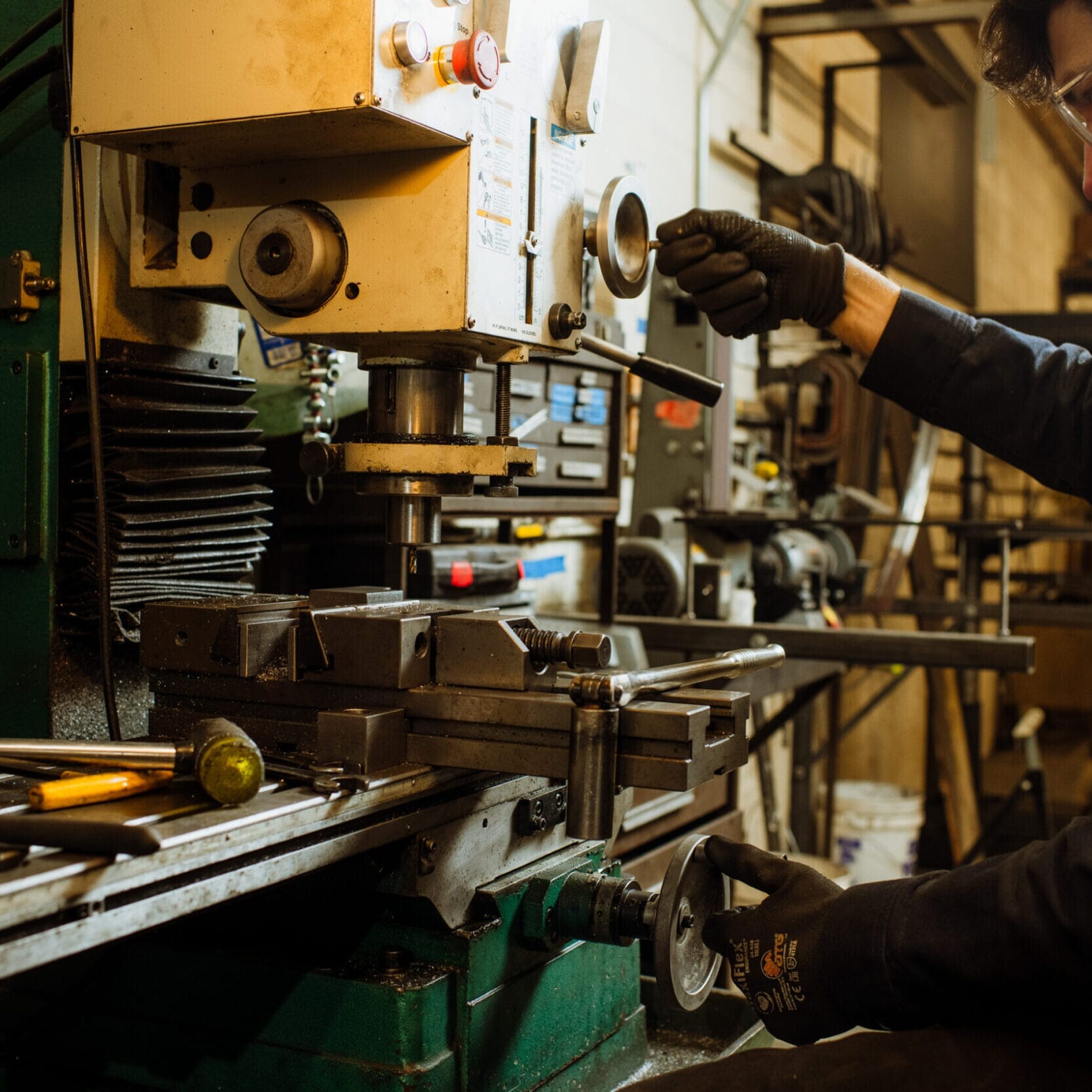 A person wearing gloves and safety glasses operates a milling machine in Dyna Metal Shop Seattle, adjusting the controls and positioning metal components for machining.