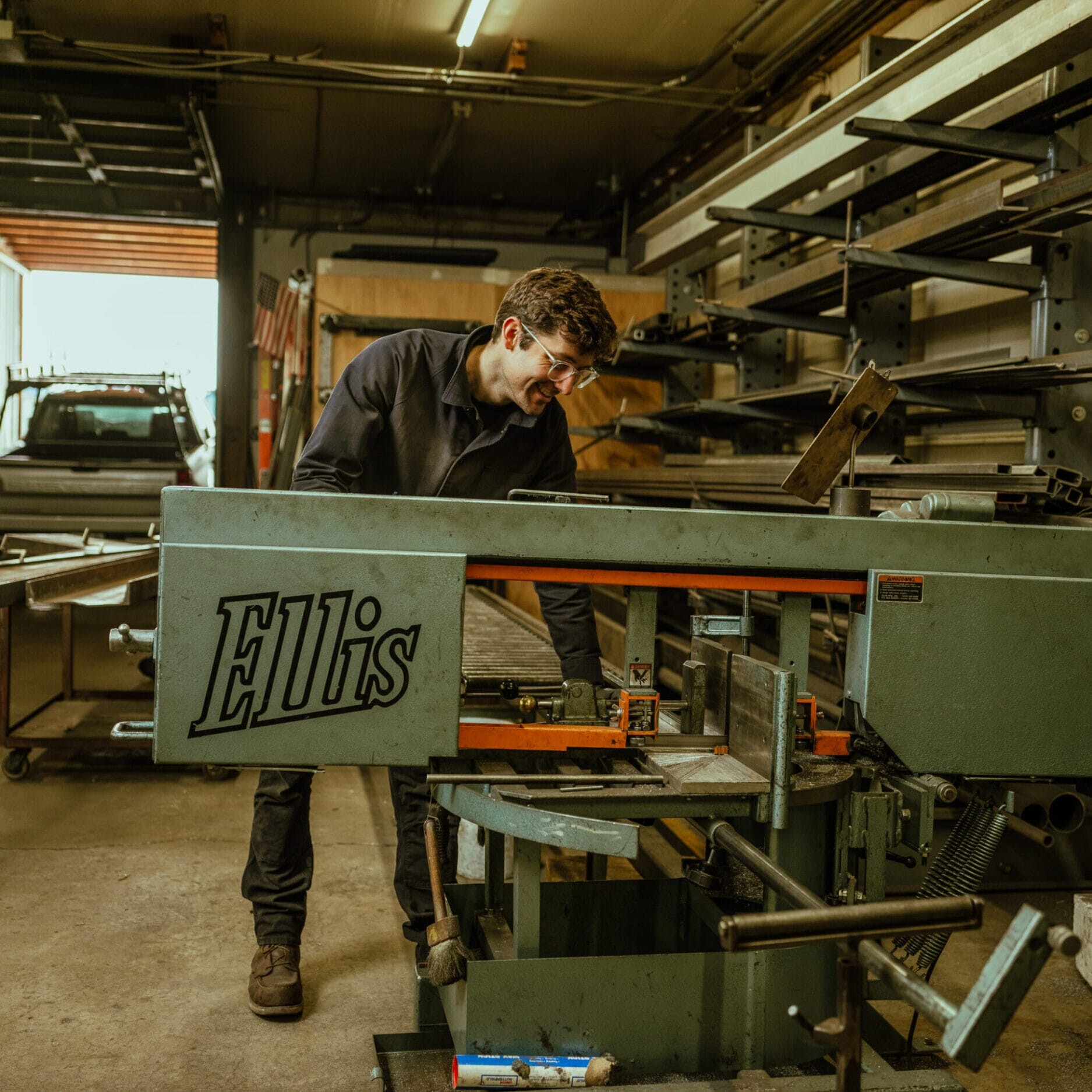 A person operates an Ellis industrial cutting machine in the Dyna Metal Shop Seattle workshop, with shelves and a parked vehicle visible in the background.