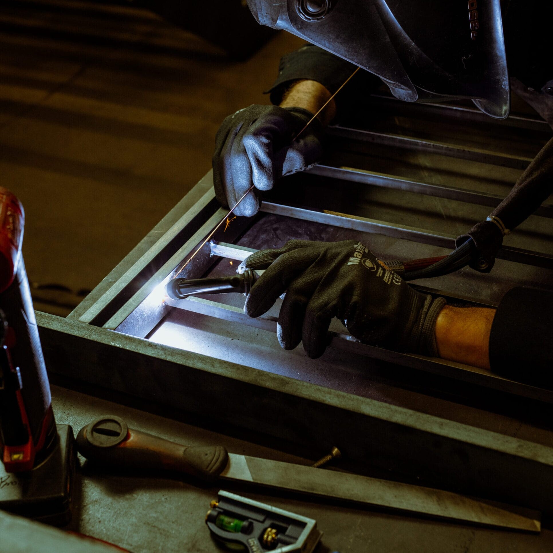 A person wearing gloves and a welding helmet uses a welding torch on a metal frame at Dyna Builders Metal Shop, with various tools visible on the workbench nearby.