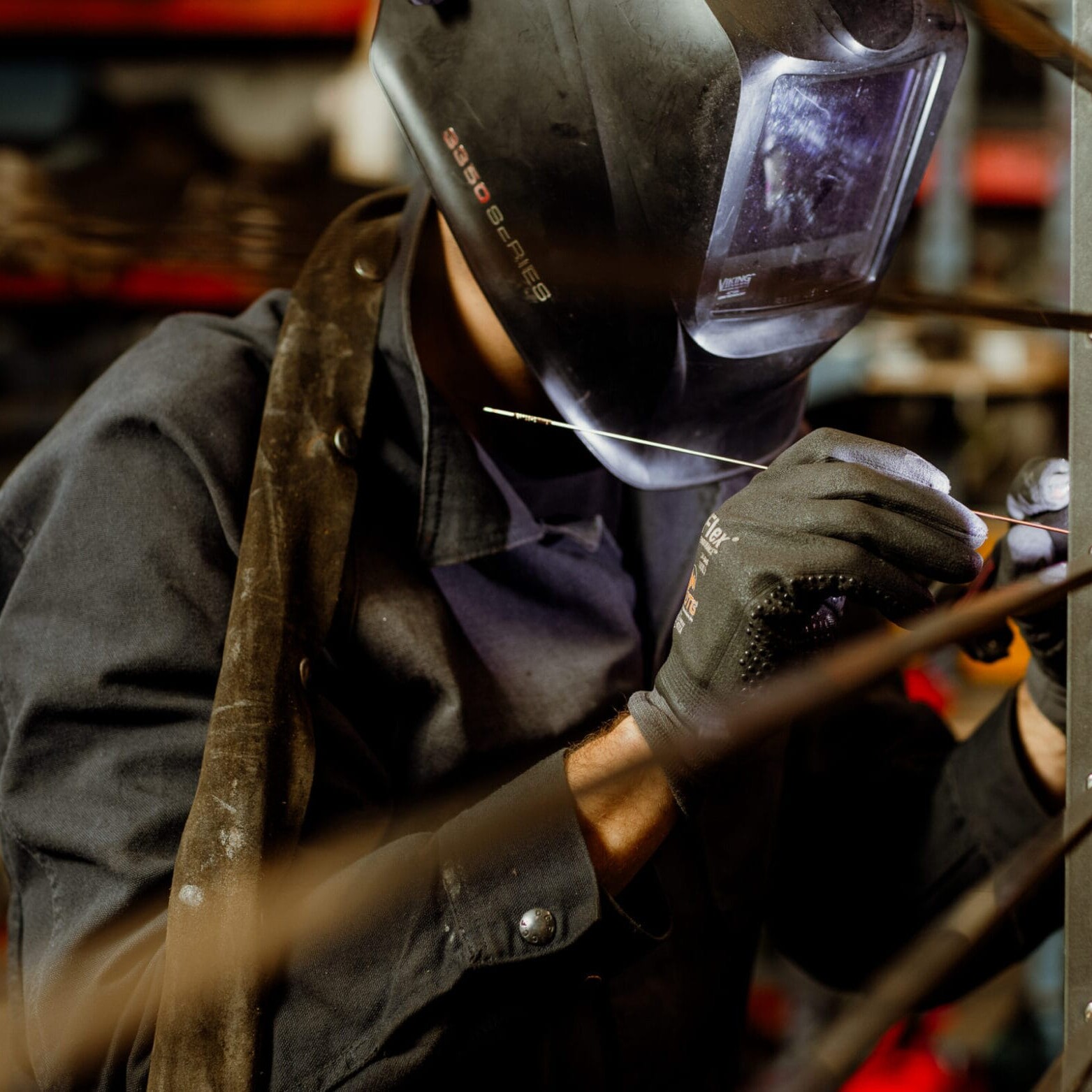 A person wearing a welding helmet and gloves is welding metal rods together in the Dyna Metal Shop Seattle workshop.