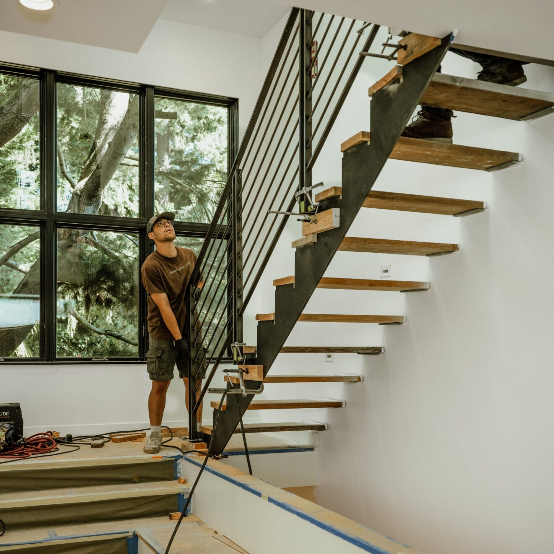 A person stands at the base of a modern floating staircase under construction in a house, with tools from Dyna Metal Fabricators scattered on the floor nearby.