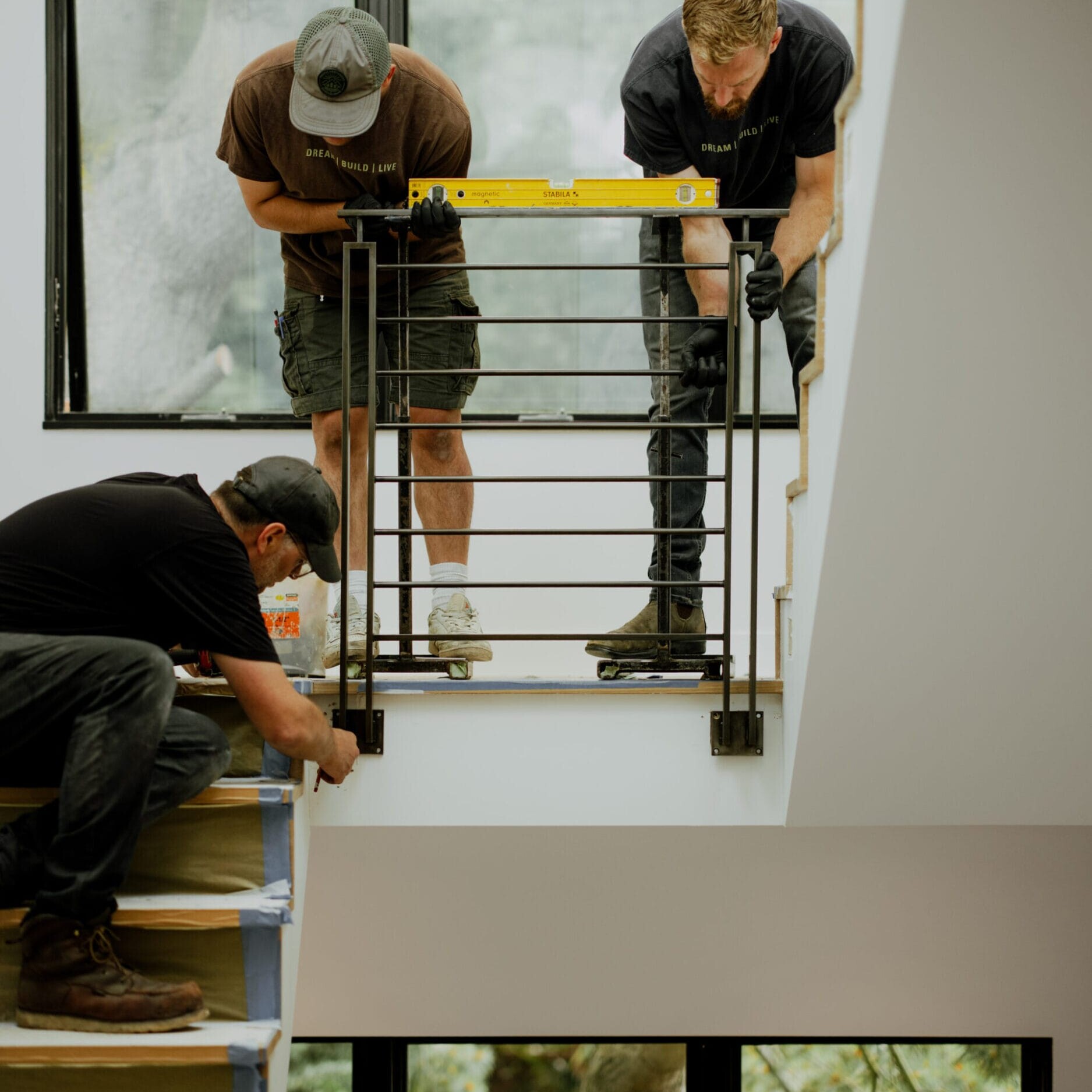 Three workers from Dyna Metal Fabricators install a metal stair railing indoors; two hold a level on top while the third secures the base at the bottom of the stairs.