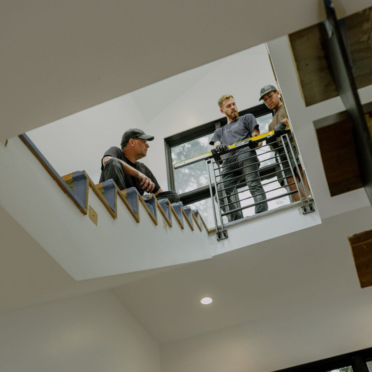 Three people stand on an upstairs landing, looking over a railing in a modern, well-lit building with wooden stairs—an example of craftsmanship by Dyna Metal Fabricators.