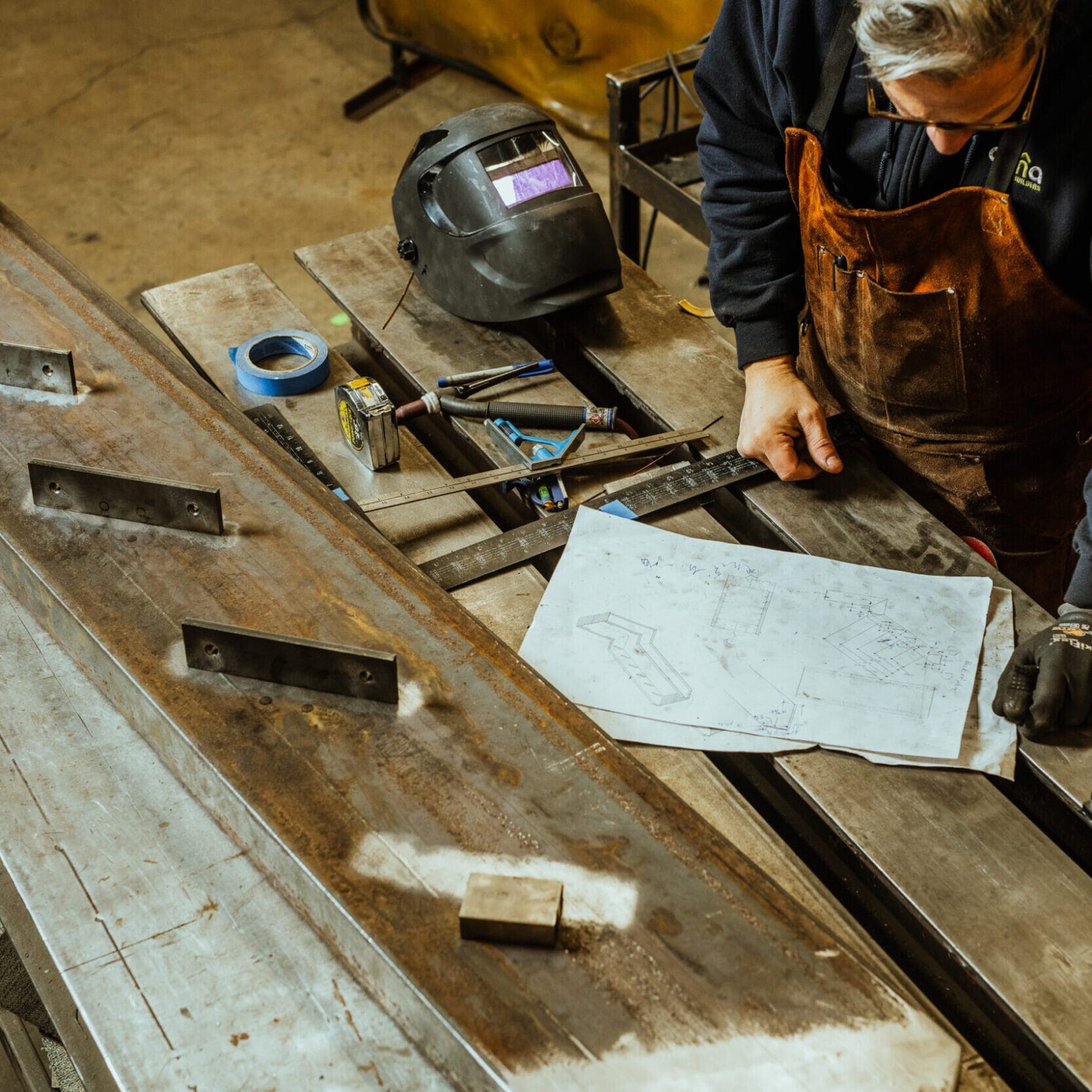 In the Dyna Metal Shop Seattle, a person in an apron examines blueprints on a workbench covered with metal parts, tools, and a welding helmet.