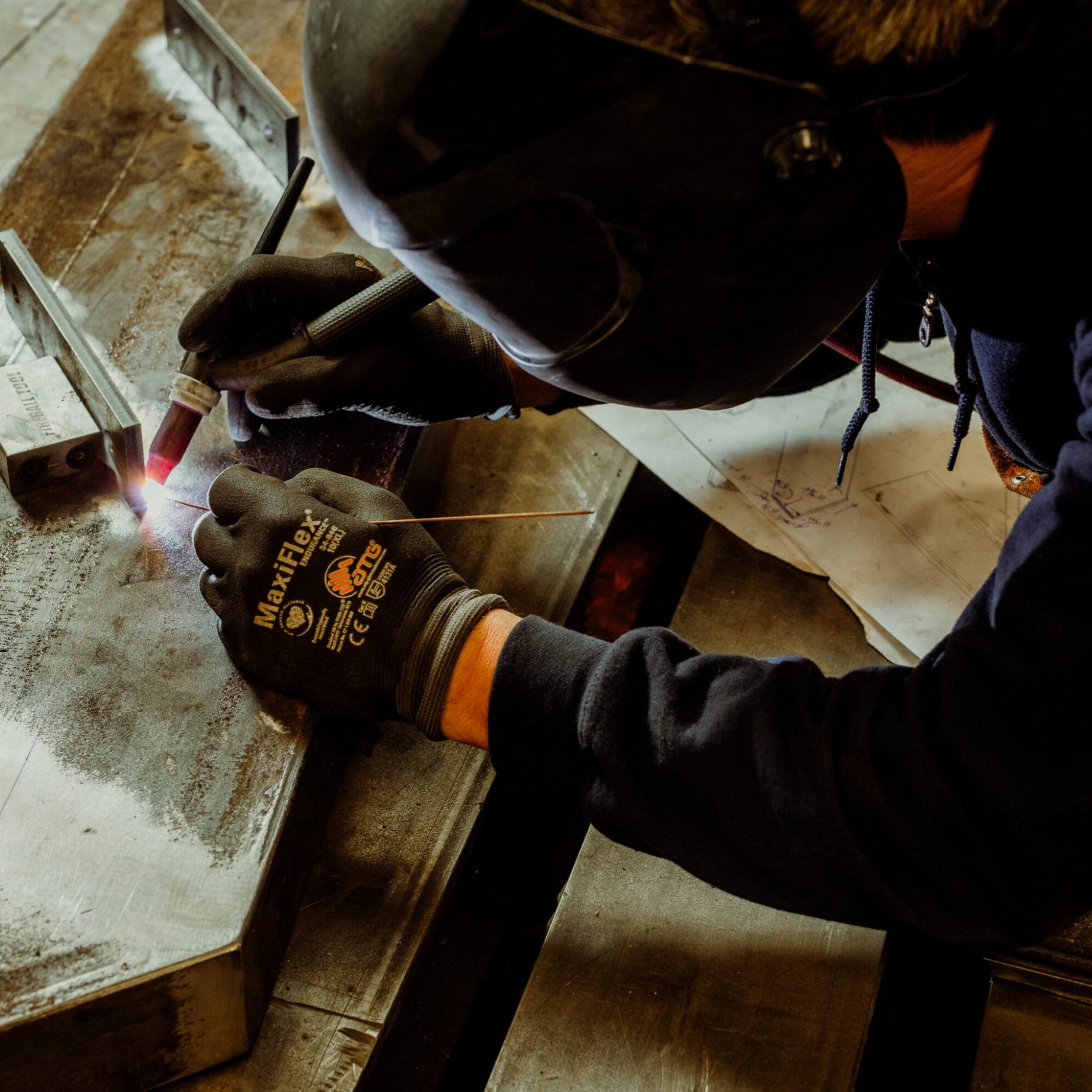 A person wearing protective gear and gloves welds metal pieces together on a workbench at Dyna Metal Fabricators.