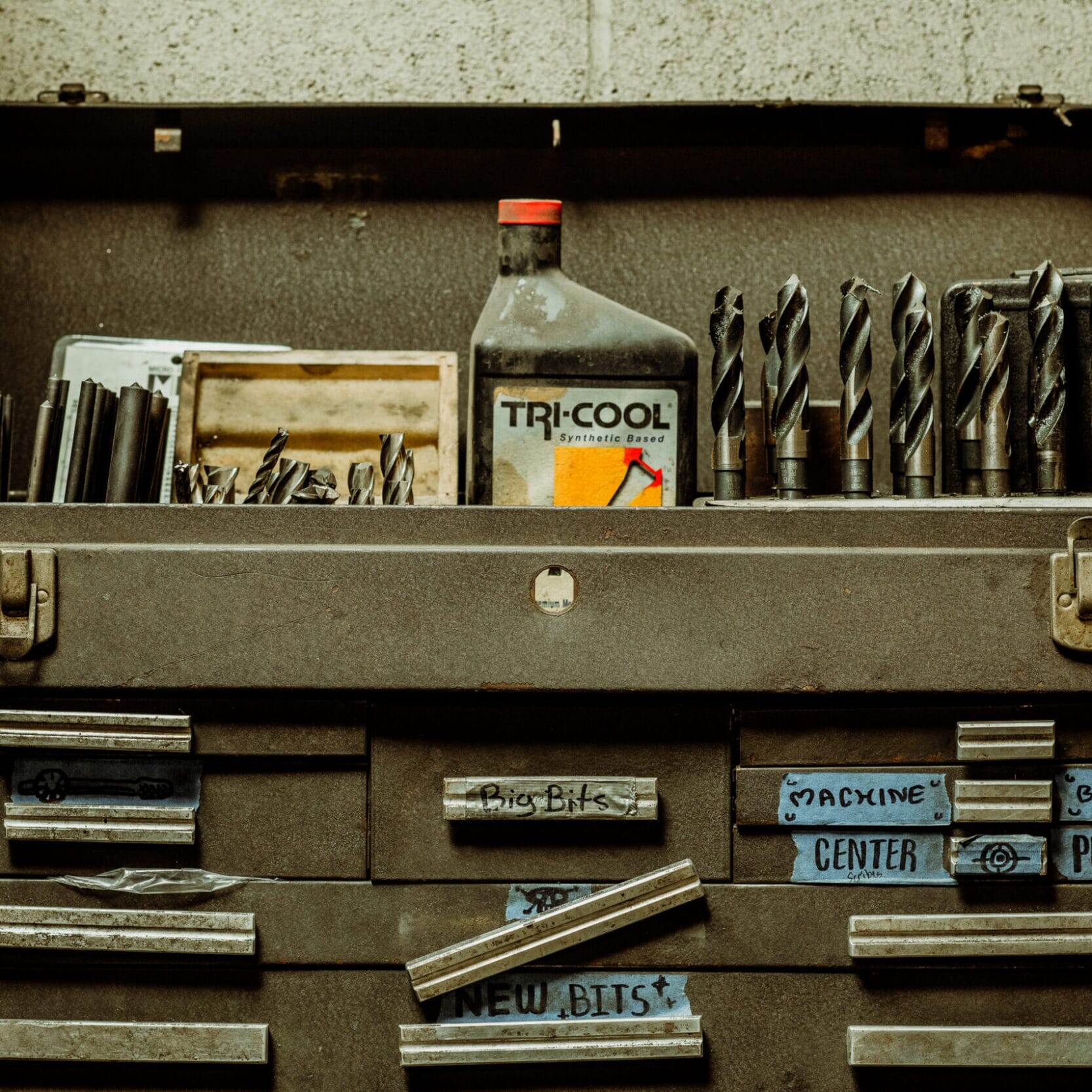 An open metal tool chest at Dyna Metal Shop Seattle holds drill bits, a Tri-Cool bottle, and labeled drawers for various tools, all set against a cinder block wall.