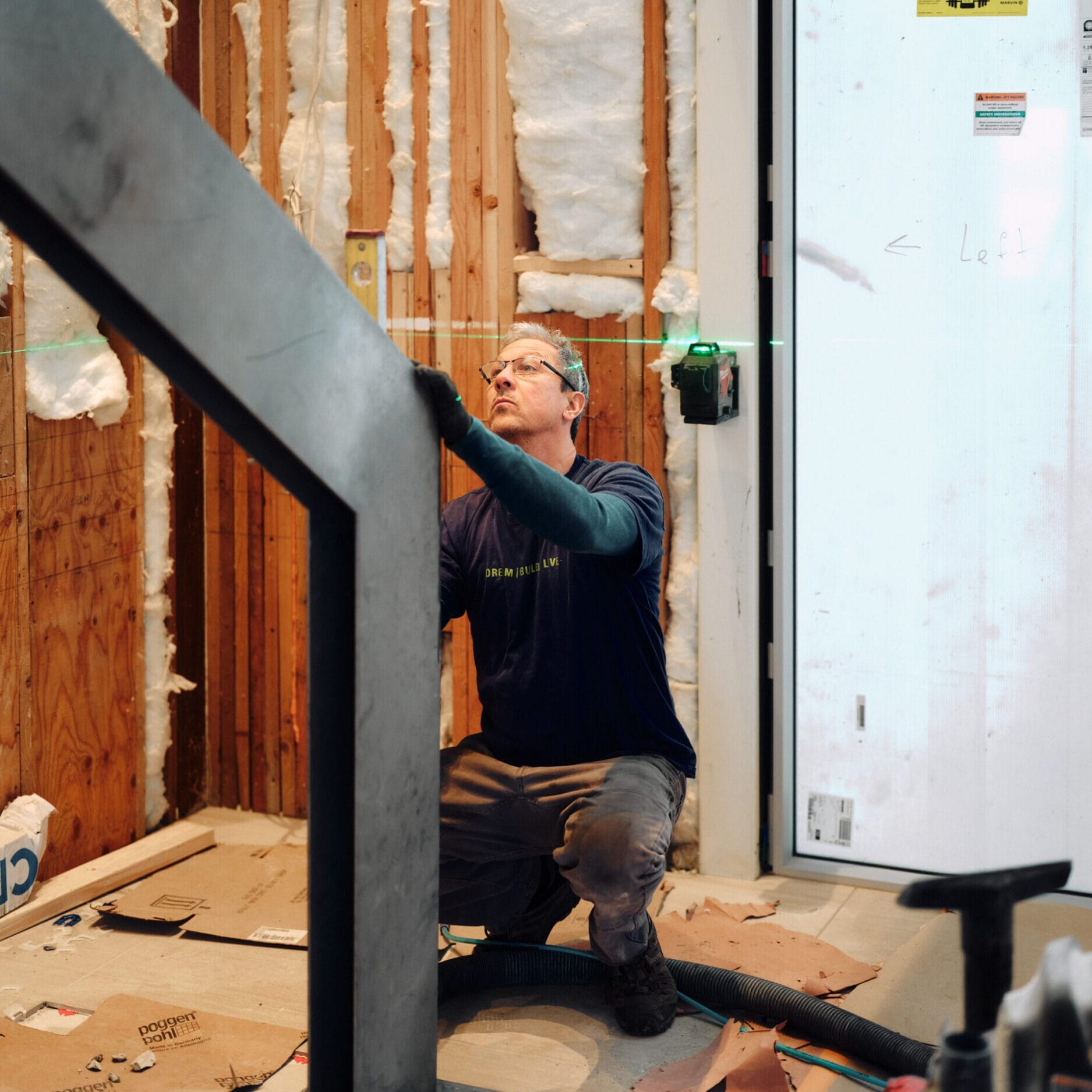A person kneels on the floor of a construction site, using a green laser level to align a metal structure near exposed insulation and an unfinished wall at Dyna Metal Fabricators.