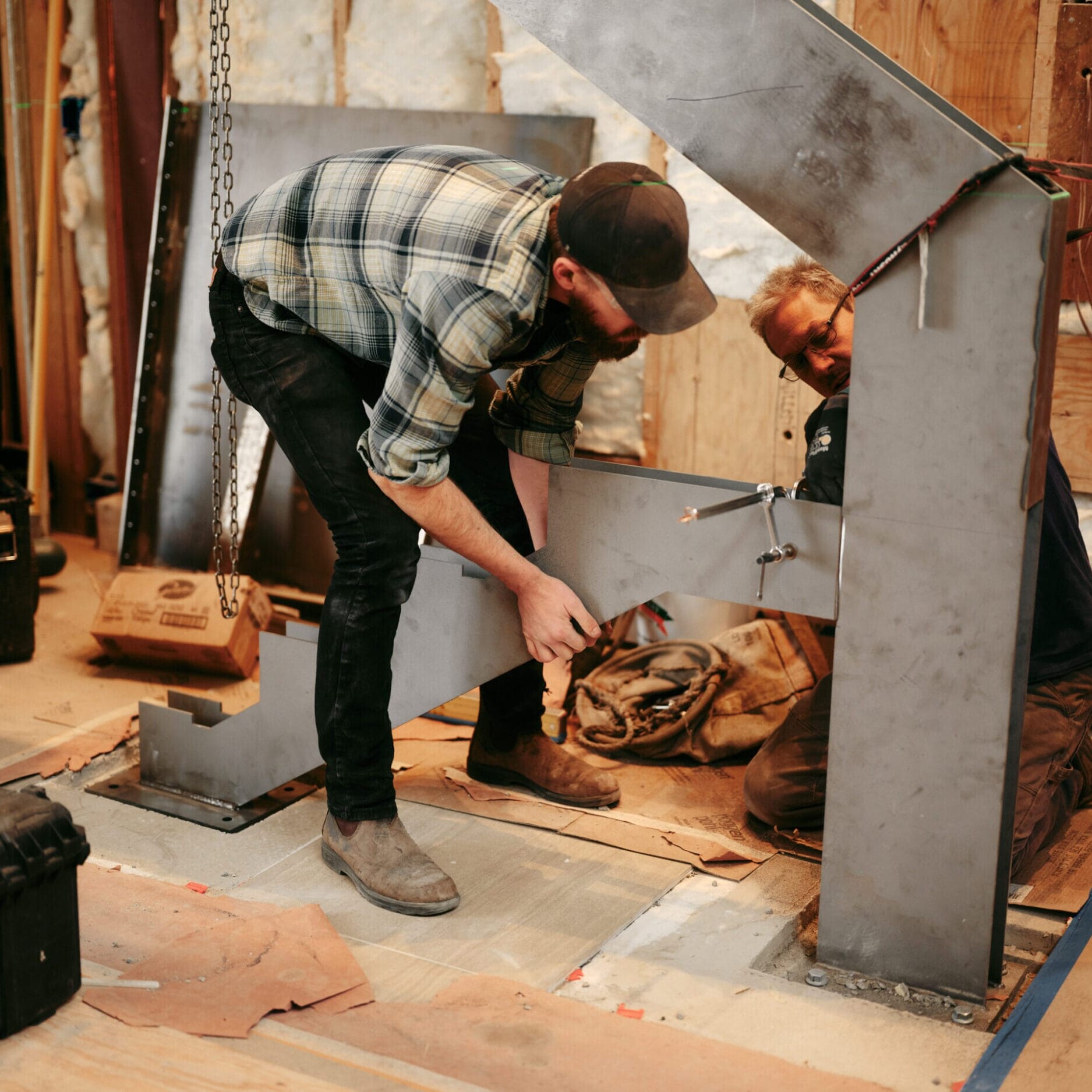 Two men install a large metal beam in the Dyna Metal Fabricators workshop, using tools and bolts, surrounded by equipment and construction materials.