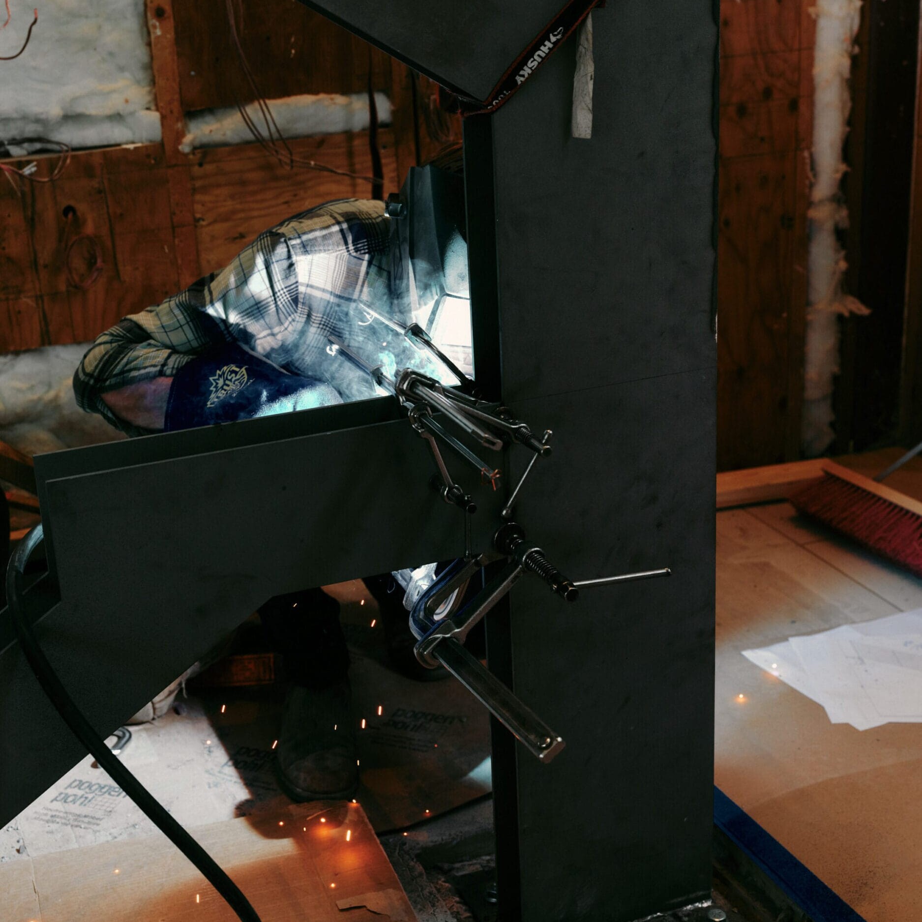 A person wearing a plaid shirt is welding metal inside a construction area with exposed insulation and wooden walls at the Dyna Metal Shop Seattle.