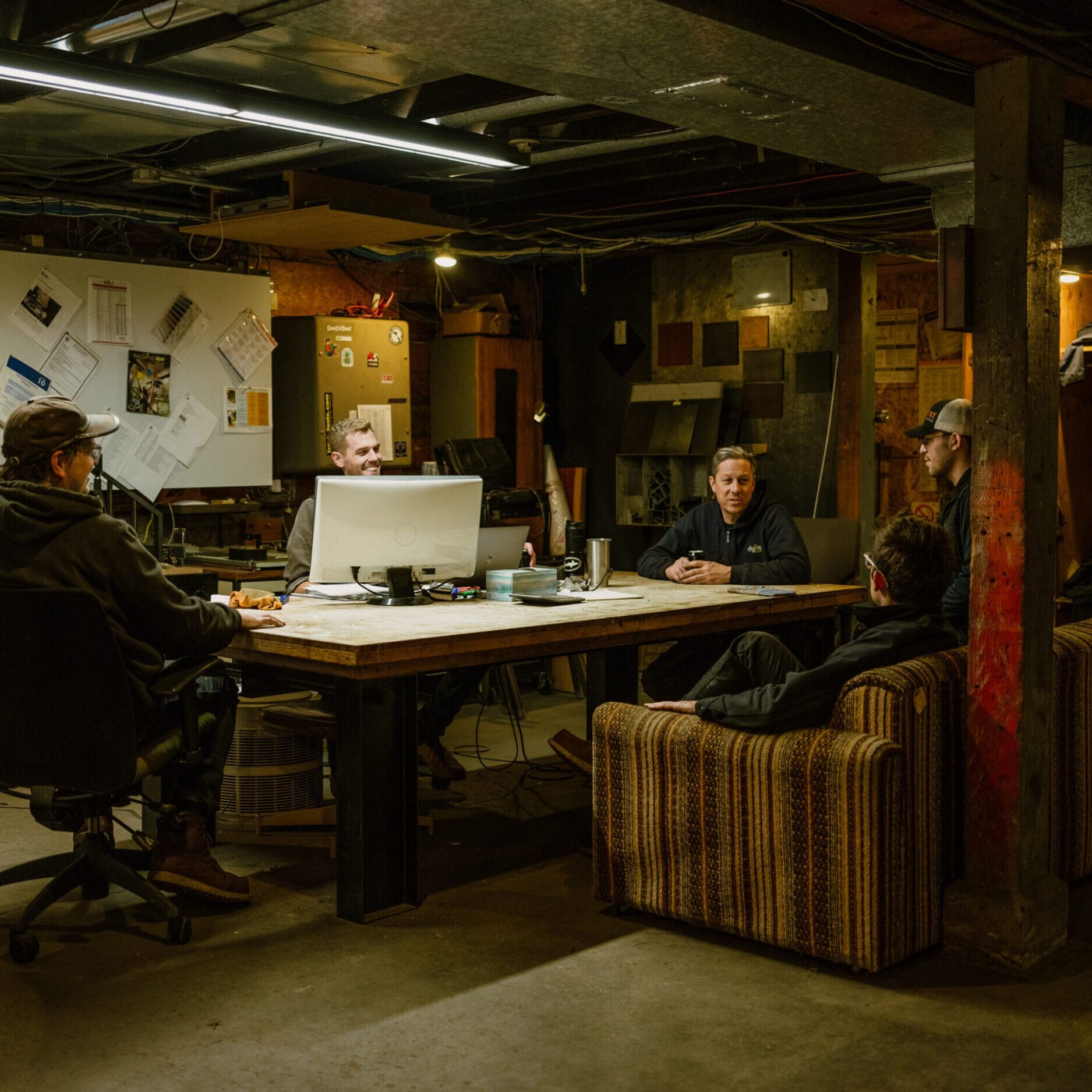 Five people sit and talk around a large table in the dimly lit, cluttered basement office of Dyna Metal Fabricators, with bulletin boards and papers covering the walls.