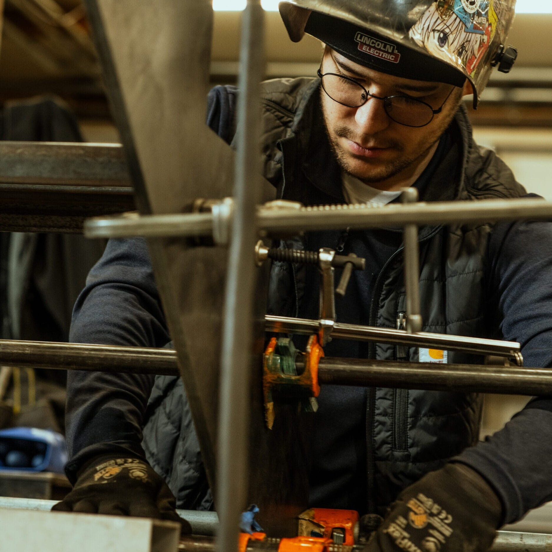 A person wearing safety gear, including gloves and a helmet, works with metal rods and clamps in the Dyna Metal Shop Seattle workshop.