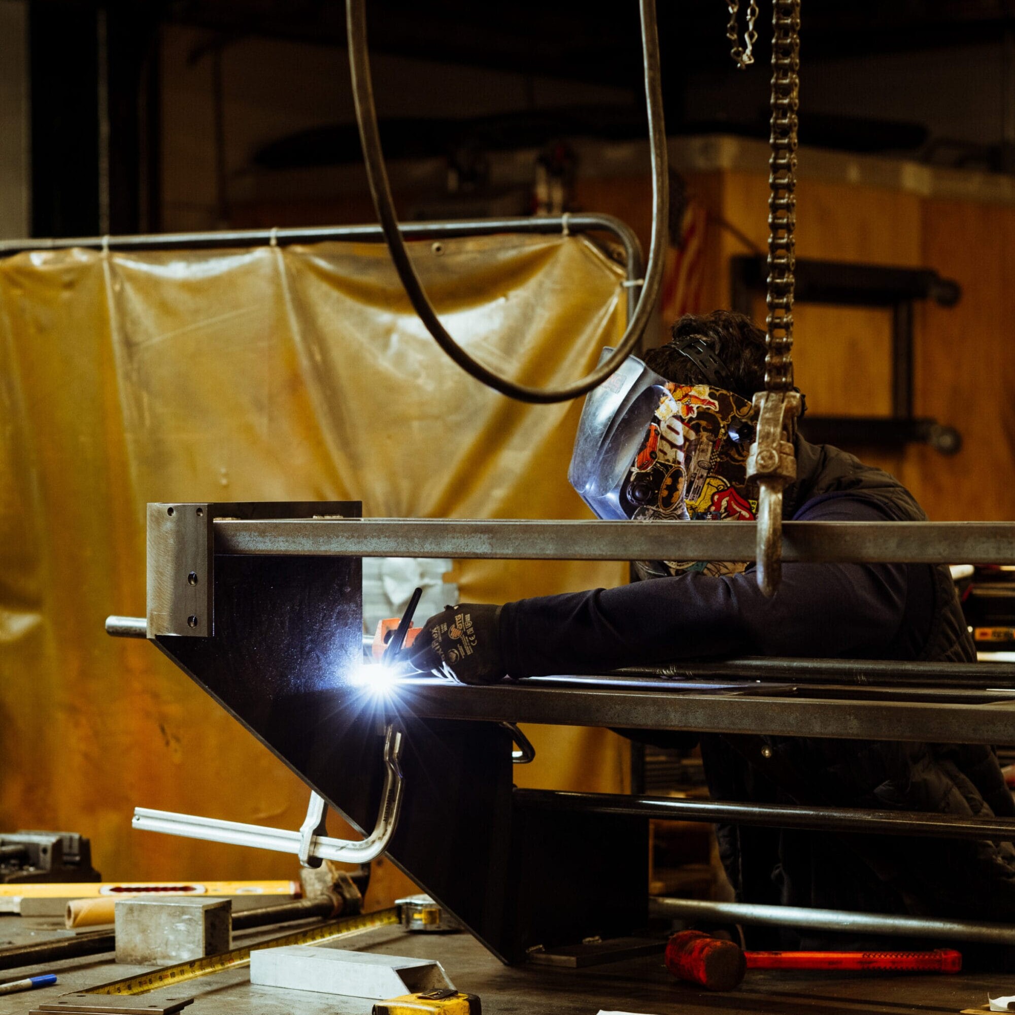 A person wearing a welding helmet works on a metal structure in the Dyna Metal Shop Seattle workshop, surrounded by tools and equipment.
