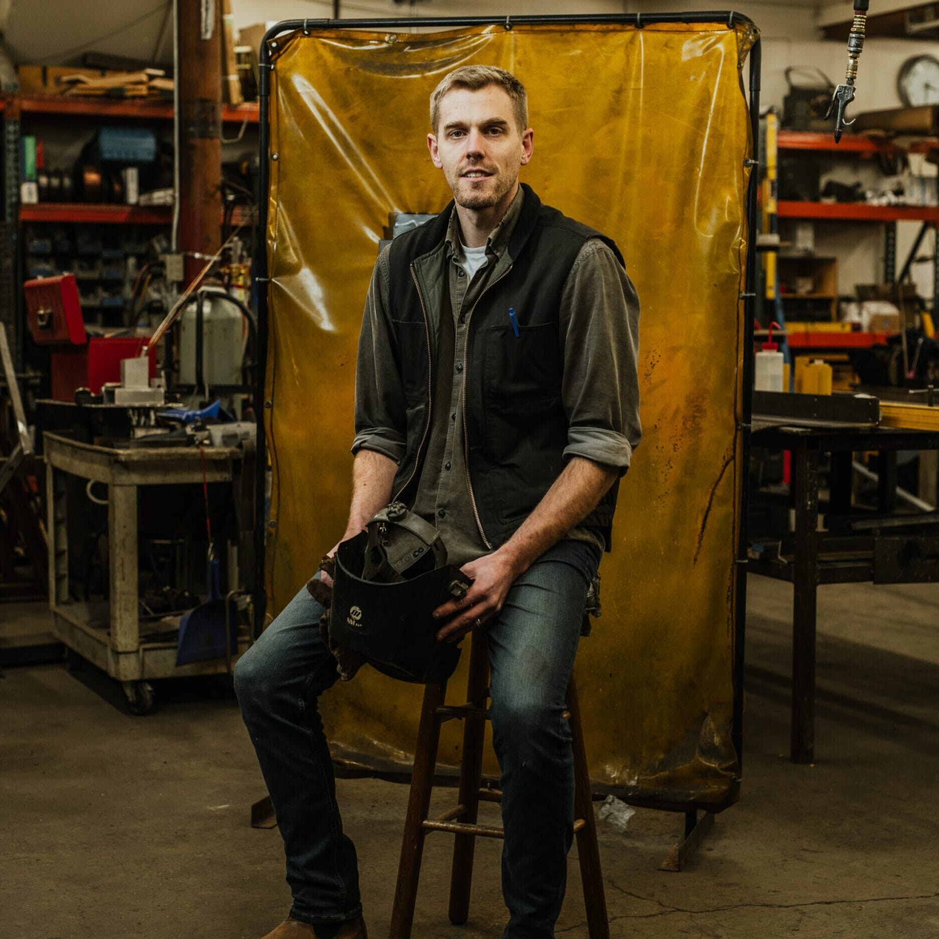 A man sits on a stool in the Dyna Metal Shop Seattle workshop, holding a welding helmet and wearing casual work clothes. Tools and equipment are visible in the background.
