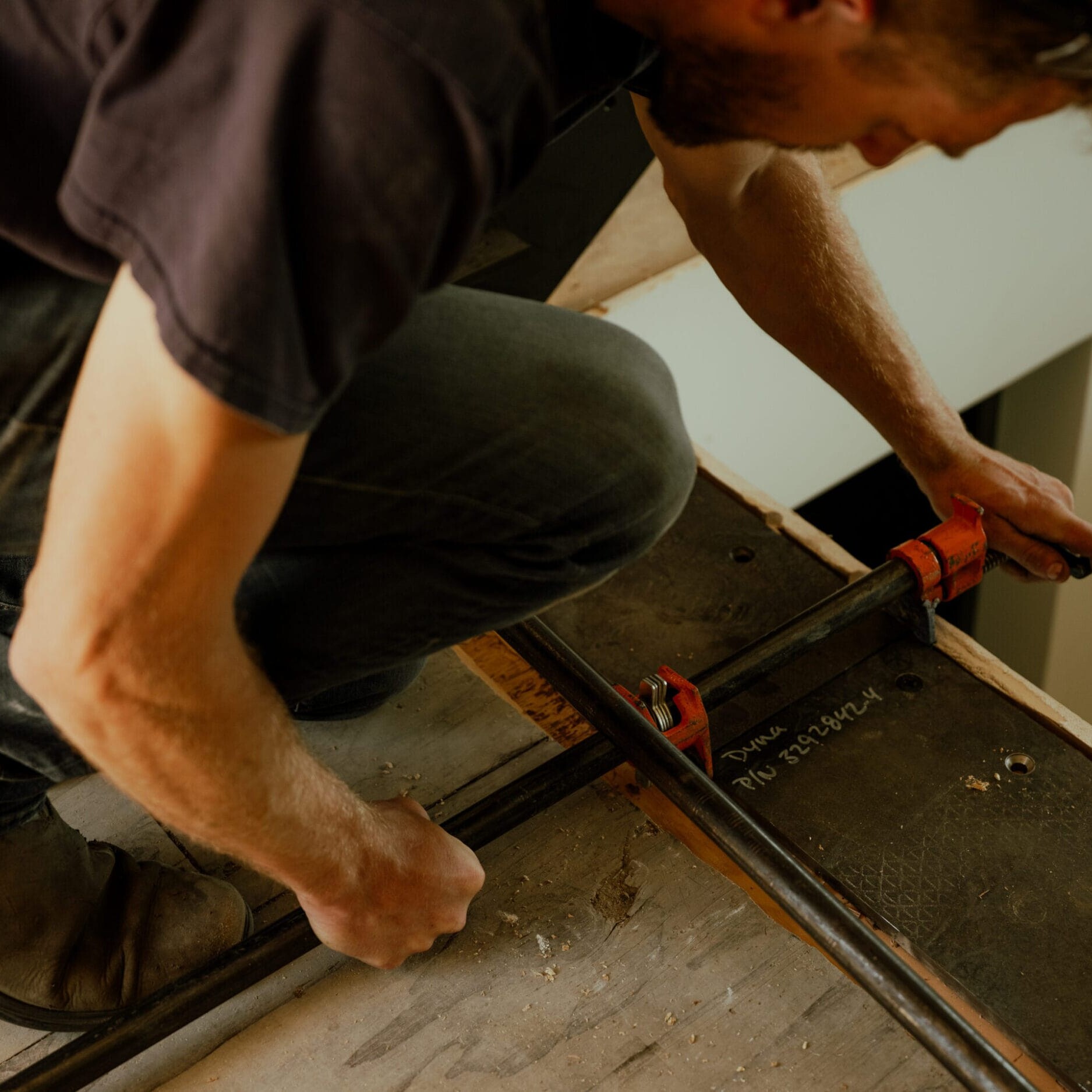 A person kneels while tightening a metal clamp on a piece of wood, working on an indoor carpentry project—showcasing the craftsmanship inspired by Dyna Metal Shop Seattle.