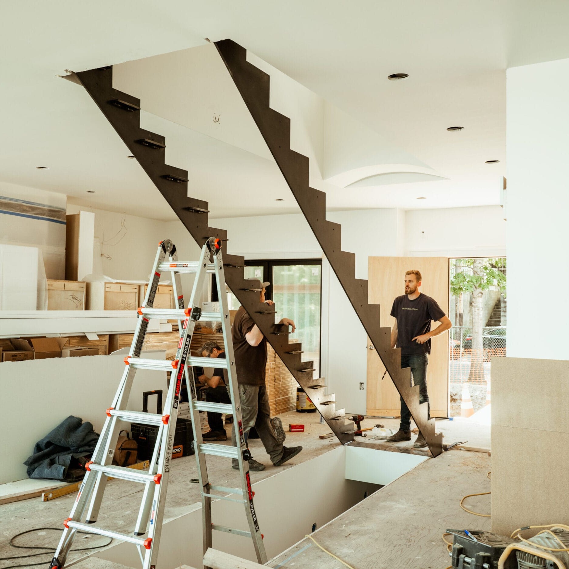 Two people stand near a partially constructed staircase in a modern, unfinished room with construction materials from Dyna Metal Shop Seattle scattered around.