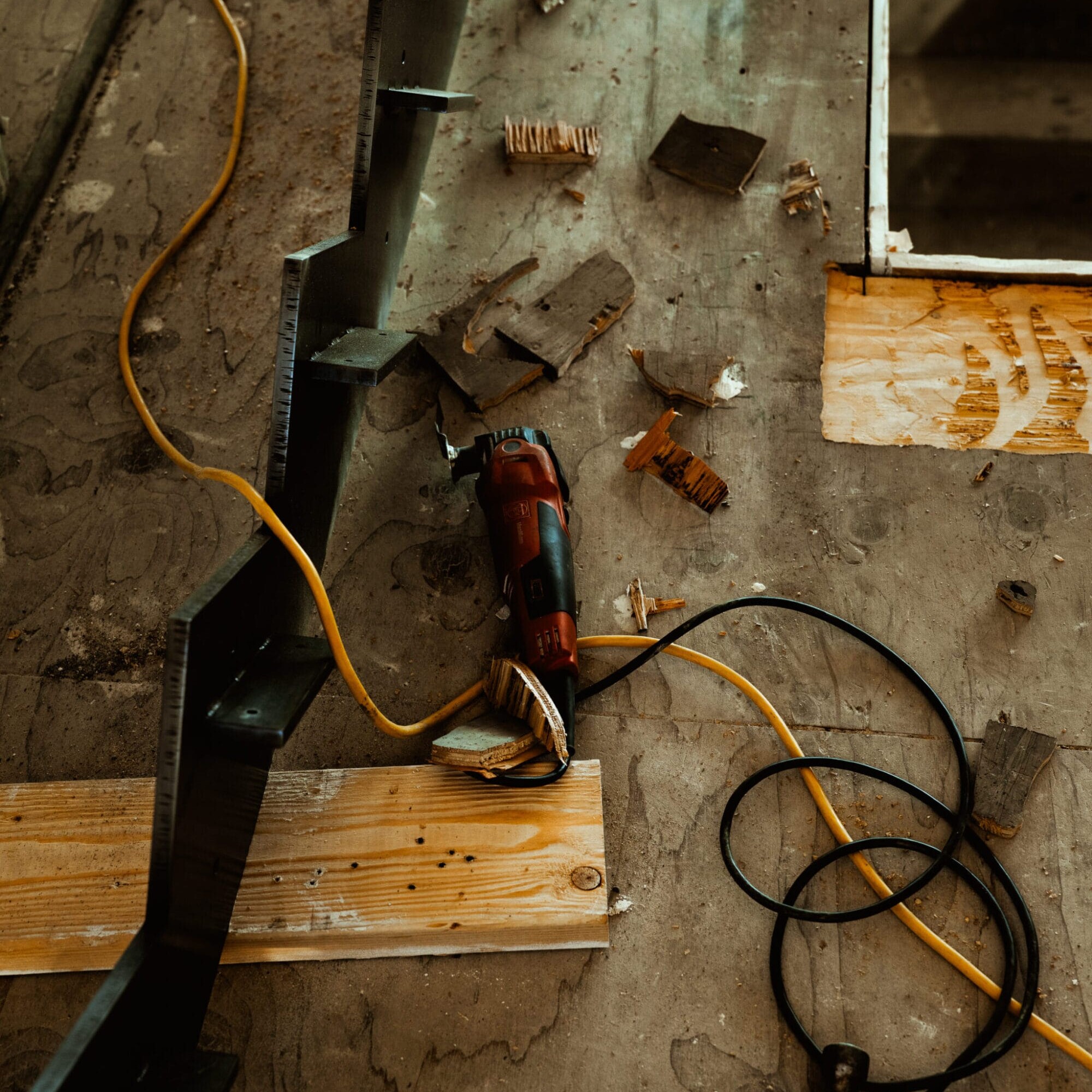 A power tool with a yellow cord lies on a dusty wooden floor surrounded by scattered wood pieces and metal parts, capturing the busy atmosphere of Dyna Metal Fabricators at work.