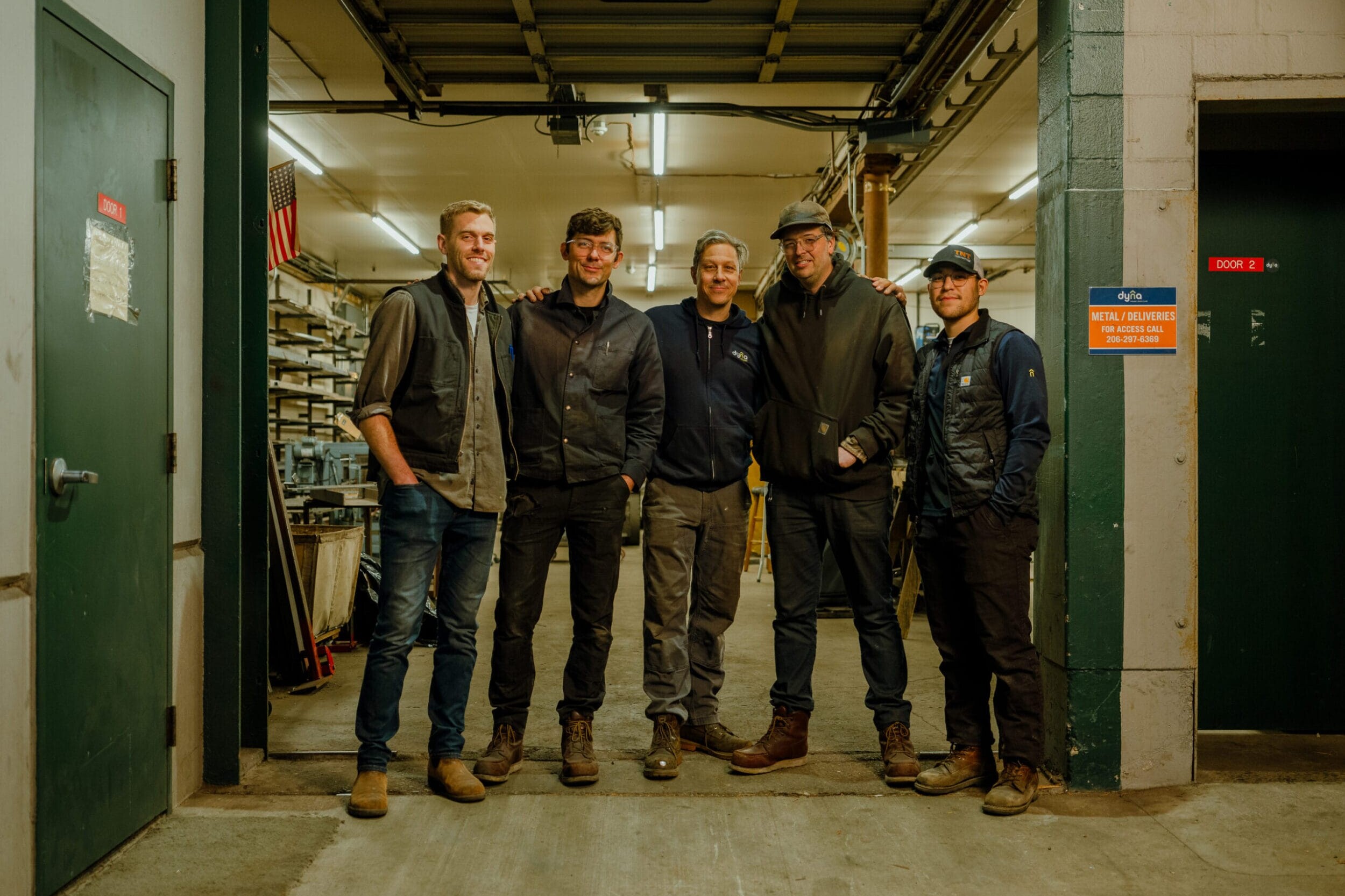 Five men stand together in the Dyna Builders Metal Shop, wearing work clothes and boots, with shelves and equipment visible in the background.